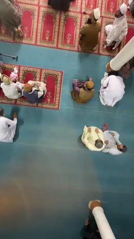 This is an overhead, slightly tilted, wide shot of a group of men praying inside what appears to be a mosque. The floor is a solid teal blue, and there are multiple red prayer rugs with gold patterns laid out on it. The men are wearing traditional Muslim attire, including white and earth-toned thobes and turbans or kufis. They are arranged in rows, facing a direction that is not visible. Some men are kneeling, some are sitting, and others appear to be in the standing or bowing positions of prayer. In the foreground, a large white pipe structure obstructs the bottom right portion of the frame. The lighting is dim, and there are no visible windows or external light sources. The overall atmosphere appears to be one of quiet reverence and communal worship.
