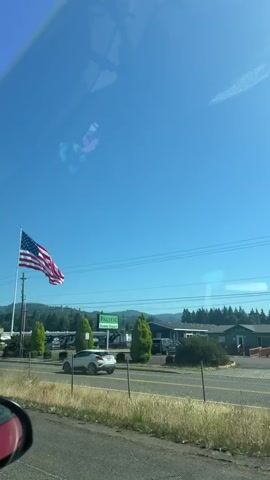 The video shows an American flag waving in the wind against a bright blue sky. The flag is on a tall flagpole to the left of the frame. In the background, there are trees, hills, and several recreational vehicles parked in a lot. A white car is driving past the RV lot. A fence is visible in the foreground. The scene appears to be outdoors during the daytime, likely on a sunny day. The sky is clear with a few faint streaks that could be contrails. The location is identified as Woodland, United States of America. There is a green sign visible in the background that says "PACIFIC HOME DEALERS".