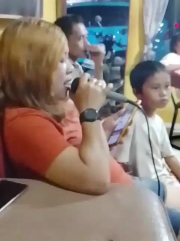 A woman with blonde hair and an orange shirt is holding a microphone and a smartphone. She appears to be singing. To her left, a man is looking away from the camera, with his hand near his face. To her right, a child in a white shirt is looking away from the camera. They are sitting on a couch in what appears to be an indoor setting. A table is in the foreground, and a smartphone is on the table. There is a poster of a motorcycle on the wall in the background.