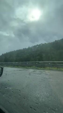 The video shows a view from a car driving on a wet, cracked road. The road is bordered by a metal guardrail and a grassy embankment. Beyond the embankment is a dense, green forest covering a sloping hill. The sky is overcast with the sun faintly visible through the clouds. The scene appears to be a natural landscape, possibly a mountainous or hilly region, with signs of recent or ongoing rain.