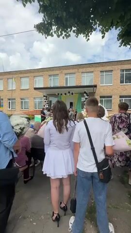 The video shows a group of people, mostly teenagers, gathered outside a brick building with a banner hanging in front of the entrance. The building appears to be a school. Many of the teenagers are dressed in formal attire, with some wearing white dresses and others in suits. They are holding bouquets of flowers, suggesting a celebratory event such as a graduation or end-of-year ceremony. Black and yellow balloons are visible, decorating the entrance. The weather appears to be overcast, with some trees visible in the foreground, casting shadows. The people are facing the building, and some are holding hands. The overall atmosphere seems to be one of anticipation and excitement.