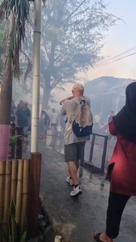 The video depicts the aftermath of a fire. The remains of a building are visible, significantly damaged, with the roof collapsed and debris scattered around. Smoke fills the air, obscuring some of the scene. Several people are present in the area, walking around the wreckage. Some individuals appear to be assessing the damage or possibly searching through the debris. A sign that reads "CAMEL FAST" is partially visible among the wreckage. The ground is wet, and there are some remnants of structures, like fences, still standing. Palm trees are visible in the foreground, adding to the overall setting.