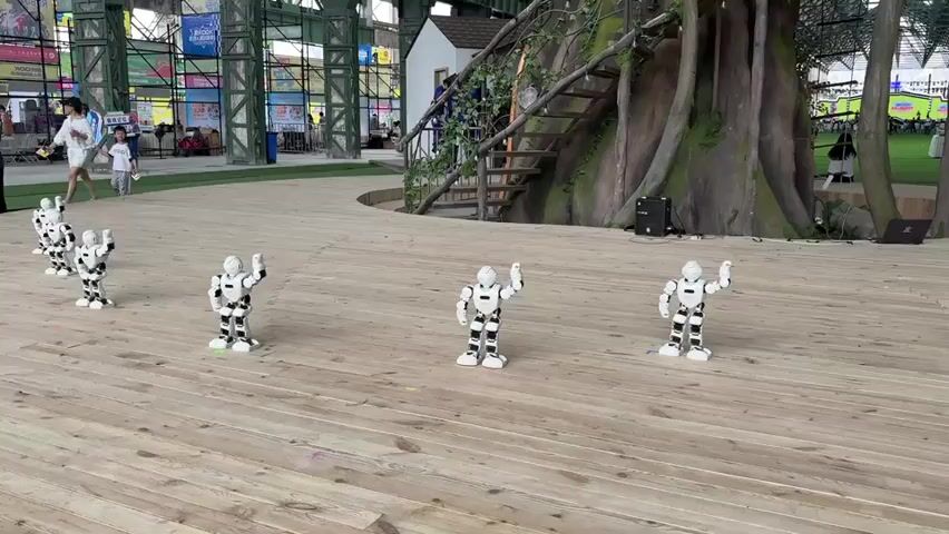 Four small white and black robots are dancing in a line on a wooden platform. Three robots are in focus in the foreground and another robot is slightly further back. A group of five robots are in the background, moving in unison. The robots appear to be performing a choreographed dance. In the background, there is a large artificial tree with a wooden staircase leading up to a small house. There are also several people visible in the background, some of whom are watching the robots. The environment appears to be an outdoor exhibition or event space, with metal structures and colorful banners visible in the distance.