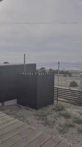 A static shot captures a coastal scene under an overcast sky. In the foreground, a wooden deck is visible, leading towards a dark, modern fence enclosing a patch of sandy ground with sparse vegetation. Beyond the fence, a broader landscape unfolds, featuring a body of water with a distant shoreline. Several white, boxy structures, possibly caravans or small buildings, are scattered across the terrain. Utility poles and power lines stretch across the middle ground, hinting at human habitation. The overall impression is one of a quiet, perhaps undeveloped, coastal area.