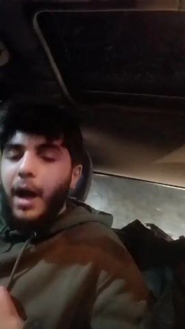 Here's a description of the video:

A young man with dark hair and a beard is sitting in the driver's seat of a car. He is wearing a dark-colored hoodie. The car has a sunroof visible at the top of the frame. It appears to be nighttime, based on the lighting inside and outside the car. The man is looking up, smiling, and singing or speaking. The video is shot from a low angle, likely by a camera positioned near the center console of the car.