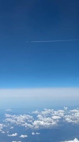 The video captures an aerial view from an airplane window. The sky is a clear, vibrant blue, with a faint haze near the horizon. A long, thin contrail stretches horizontally across the upper portion of the frame, indicating a distant aircraft. Below this, the horizon is blurred by atmospheric conditions. The lower half of the video displays a layer of fluffy white clouds, with glimpses of snow-capped mountains peeking through them. The perspective suggests the viewer is flying at a high altitude, looking down upon the landscape and sky. The camera appears to be slightly tilted, and there are moments of slight blurriness.