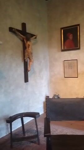 The video shows an indoor room with antique furnishings. On the left wall, a large wooden crucifix with a figure of Jesus is mounted. To the right of the crucifix, there is a simple wooden stool. In the center of the room, a heavy, dark leather armchair with studded detailing is positioned. Behind the armchair, a wooden desk with carved doors and multiple drawers is visible. On the desk, there are two books stacked, a small candle in a holder, and two decorative glass jars. Above the desk, three framed portraits hang on the wall. The first portrait on the left is partially visible, showing a person in a red hat and robe. The other two portraits are also framed and hanging. Further to the right, a smaller wooden cabinet with drawers is visible, topped with two candlestick holders. A portion of a window with multiple panes is seen in the background. The floor is covered with an ornate rug. The walls are painted with a textured finish in muted tones. The lighting is dim, suggesting ambient light from the window and possibly interior lighting. The overall impression is of a historical or museum-like setting.