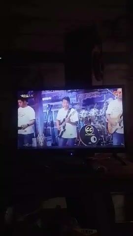 A television screen displays a live performance of a band. Two men in white t-shirts with a logo are playing keyboards in the foreground. Behind them, other band members can be seen playing instruments, including a guitar and drums. The background is dimly lit with a stage setup. The video is viewed from a low angle, indicating it's likely captured from a seated position.