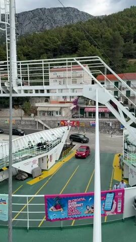 A ferry deck is visible with several cars parked on it. A red minivan is positioned towards the front of the deck. A dark colored sedan is further back. A person in a yellow vest is standing near the cars. A banner with text and images is attached to the railing. In the background, buildings and green trees are visible on a hillside. A steep, rocky mountain rises behind the trees. The sky is cloudy.