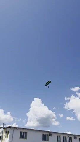 A wide-angle, low-angle shot shows a skydiver descending under a green and black parachute in a bright, clear blue sky dotted with fluffy white clouds. The skydiver is a small figure beneath the large canopy. Below, a white building with yellow-framed windows stretches across the lower part of the frame. The sky is predominantly clear, with the clouds concentrated near the horizon.