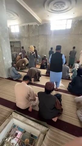 A group of men and one woman are gathered inside a room, appearing to be praying. The room has bare concrete walls with some water damage and a white ceiling with a decorative molding. There are several windows with sunlight streaming through them. The floor is covered with striped mats. Most of the people are wearing traditional clothing such as tunics, vests, and head coverings. Some individuals are kneeling or prostrating on the mats, while others are seated. In the foreground, on the left, there is an opening in the floor revealing another group of people, also seemingly praying, with a more ornate ceiling visible below. The overall atmosphere suggests a religious gathering.