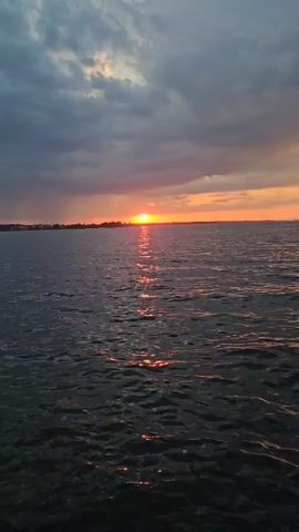 The video depicts a sunset over the water. The sun is positioned on the horizon, glowing orange and reflecting on the dark, rippling surface of the water. Dark clouds fill much of the sky, while an orange glow permeates the horizon.  A faint land mass appears on the horizon beneath the setting sun.