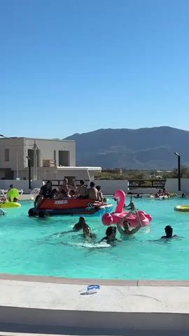 A group of people are enjoying a sunny day at a swimming pool. In the foreground, several individuals are swimming and interacting. To the left, a group of people are seated on an inflatable raft that appears to be a boat. A large pink flamingo inflatable is also present in the pool. In the background, more people are seen in the water, and some are relaxing on inflatables. The pool has clear, turquoise water and is surrounded by a concrete deck. Beyond the pool, there is a modern building and a lush green landscape leading to mountains under a clear blue sky. The overall atmosphere is one of recreation and enjoyment.