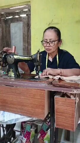 A middle-aged Asian woman wearing glasses and a dark blue shirt with a yellow measuring tape around her neck is sewing with a vintage black sewing machine. The sewing machine is placed on a wooden table. The woman is focused on her work, her hands guiding the fabric under the needle. The background consists of a pale yellow wall and a wooden door. Underneath the table, there are some items, including a colorful patterned fabric or material. The scene appears to be an indoor setting, possibly a home or a small workshop.