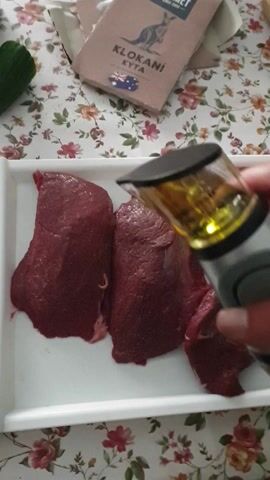 The video shows a close-up, overhead view of a person's hand pouring oil from a clear bottle with measurement markings onto several pieces of raw red meat on a white rectangular tray. The meat appears to be beef or venison. The tray is placed on a table covered with a floral tablecloth. To the left of the tray, a box with text and a picture of a kangaroo is visible. A green zucchini is also partially visible in the bottom left corner. The lighting is natural, suggesting an indoor setting. The hand is holding the bottle and tilting it to allow the oil to flow onto the meat. The action is focused on seasoning the meat.