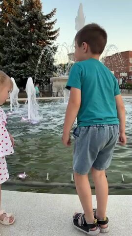 Two children are dancing in front of a fountain. A boy in a teal t-shirt and denim shorts is dancing enthusiastically, while a younger girl in a white and red polka dot dress stands beside him. The fountain has multiple water jets and is illuminated with colored lights. A tall evergreen tree is visible behind the fountain on the left. The sky is clear and appears to be late afternoon or early evening. Another child, mostly out of frame, is visible on the right side of the screen.