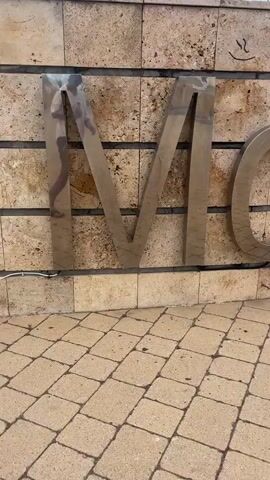 The video shows a close-up view of large, metallic letters mounted on a wall. The wall appears to be made of natural stone tiles in varying shades of beige and brown. Below the wall, there is a tiled walkway composed of interlocking rectangular pavers in a light tan color, arranged in a diagonal pattern. The metallic letters form words, and the camera pans across them. In the first few seconds, the letters "M," "O," and parts of another letter are visible. As the camera continues to move, the letters "O," "H," "L," "D," "E," and "C" are revealed, suggesting the word "MOHLDEC" or a similar combination. The lighting suggests it is daytime, possibly outdoors, given the reflection on the metallic letters. There are no people visible in the video.