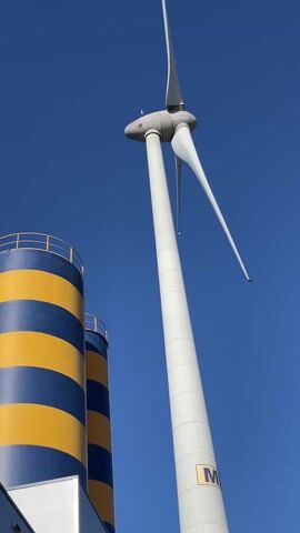 The video shows a low-angle view of a white wind turbine under a clear blue sky. The turbine is stationary, with its three blades angled irregularly. Beside it are two cylindrical tanks with alternating horizontal stripes of blue and yellow, each tank topped with a metal railing. At the bottom of the frame, a small portion of a white building is visible. There is no visible movement or interaction.