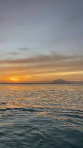 This video captures a tranquil ocean scene at sunset. The sun is visible as a bright orange orb on the left horizon, casting a warm glow across the sky and reflecting on the water. The sky is a gradient of soft blues, grays, and oranges, with wispy clouds streaking across. The ocean's surface is calm, with gentle ripples that catch the light. In the distance, a silhouetted landmass, possibly an island or mountain range, is faintly visible against the horizon. The camera appears to be positioned low, close to the water, giving a sense of immersion in the seascape. The overall mood is peaceful and serene.