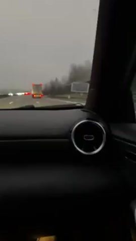 The video shows a view from inside a car driving on a highway on a foggy day. The dashboard of the car is visible, showing a digital display with information, and circular air vents. Other cars and a truck can be seen on the highway ahead. The environment outside the car appears overcast and hazy due to the fog. Bare trees line the side of the highway.