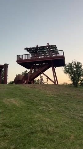 The video shows an outdoor scene with a large wooden observation tower on a grassy hill. The tower is red and has a deck at the top, accessible by stairs. Two children are visible on the stairs, one in a yellow shirt and another in a blue shirt. Further back in the park, two people are sitting in wheelchairs near some trees. The sky is clear with a hint of sunset colors. The overall environment appears to be a park or recreational area with green grass, trees, and a clear sky.