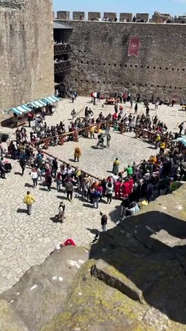 The video shows a large outdoor courtyard enclosed by stone walls, filled with a crowd of people watching two individuals engaged in a sword fight. The combatants are dressed in medieval-style armor, with the crowd surrounding the fighting area, partially enclosed by wooden barriers. Some people are taking photos or videos with their phones, while others are simply watching. There are tents and tables set up around the perimeter, suggesting a festival or fair. The ground is paved with light-colored stone tiles arranged in a random pattern. A red banner with a symbol hangs on one of the stone walls. The video appears to be shot from an elevated position.