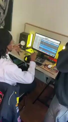 The video shows an indoor scene of what seems to be a small home recording studio. Two people are visible, one seated at a desk facing a computer screen, with studio monitors on either side. The computer shows a music production interface. The person sitting at the desk wears a white shirt and a black beanie. Another person, wearing a dark hoodie, stands nearby. The camera then pans to show a small, bare room with a desk, a mirror, and some plants. The walls are white, and the floor appears to be parquet.