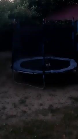 The media file captures a trampoline in a backyard setting, likely during dusk or early evening, given the dim lighting and shadows. The trampoline is enclosed with a black safety net, and its blue mat is visible. There appears to be a light-colored object, possibly a stuffed animal or a small toy, resting on the trampoline mat. The ground surrounding the trampoline is covered in dirt and some sparse vegetation. In the background, there are trees and bushes, suggesting a residential area. The overall mood is quiet and still, with no discernible activity taking place other than the object's presence on the trampoline. The time of day is implied by the low light conditions, indicating it is either dawn or dusk. There is no visible text in the media file. The location context provided is Kavetchyna, Ukraine.