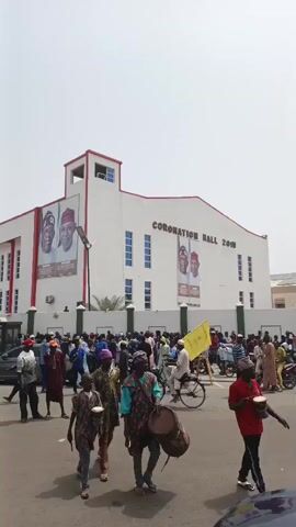 The image captures a bustling street scene in front of a large white building identified as "CORONATION HALL 2015" in Giginyu, Nigeria, during daylight hours under a clear sky. A substantial crowd of people, predominantly men, is gathered and moving in the foreground and midground. Some individuals are on motorcycles, and one person is seen with a bicycle. A person in the foreground carries two drums, suggesting a festive or ceremonial atmosphere.

Prominently displayed on the building's facade are two large vertical banners featuring portraits of political figures. One banner clearly shows "PRESIDENT" and "ALL PROGRESSIVES CONGRESS" along with the names "TINUBU SHEYTTMA". The other banner, partially visible, likely promotes another political figure for "GOVERNOR." These banners indicate a political event or rally.

Many in the crowd are dressed in traditional Nigerian attire. A bright yellow flag, possibly bearing a political slogan, is held aloft by someone in the crowd. People appear to be engaged, with some holding white bowls or cups. The scene conveys a sense of community gathering or political assembly.