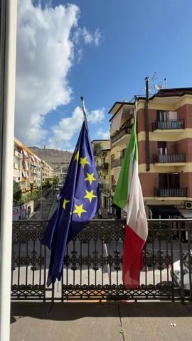 Two flags, the European Union flag (blue with a circle of yellow stars) and the Italian flag (vertical green, white, and red stripes), are prominently displayed on a balcony, hanging from poles. The flags are a central focus against a bright blue sky dotted with fluffy white clouds. Below the flags, a decorative black wrought-iron railing marks the edge of the balcony.

In the background, a residential street unfolds, lined with multi-story buildings that feature balconies and various architectural styles, common in European towns. Some buildings are painted in warm hues like yellow and orange, while others show more muted tones. A white vehicle is partially visible on the street below. In the distance, undulating hills or mountains rise, suggesting a landscape beyond the urban environment. The scene is bathed in clear sunlight, indicating daytime and good weather. There is no visible text. The location is Maddaloni, Italy.