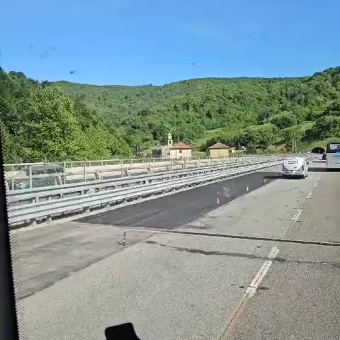 The video shows a view from a car driving along a road. In the background is a lush, green, forested mountain. A cluster of buildings, including a church with a bell tower, is visible beyond the roadside barrier.  A couple of vehicles pass in the opposite direction. The road surface appears newly paved. The sky is blue.