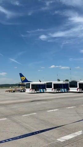 This media file captures a sunny day at an airport in Balice, Poland. The scene is dominated by an airport tarmac under a bright blue sky with scattered white clouds. In the foreground, a multi-segment airport shuttle bus, white with blue stripes and the "LS" logo, is parked. The bus is positioned on the tarmac, likely awaiting passengers or having just dropped them off.

Behind the bus, the tail fin of a Ryanair airplane is visible, clearly identifiable by its yellow logo and blue lettering. The presence of the airplane and the bus strongly indicates an active airport environment. Other airport service vehicles, such as a yellow baggage tractor, are also present near the airplane. The overall impression is one of a typical operational day at an airport.

The time of day appears to be midday, given the bright sunlight and clear skies, suggesting pleasant weather. The setting is clearly an outdoor airport environment, characterized by the vast expanse of the tarmac and the presence of aircraft and airport vehicles. While no specific emotions are overtly displayed, the scene conveys a sense of routine and functionality associated with air travel.

Visible text includes the "LS" logo on the side of the bus and the Ryanair branding on the airplane's tail. Additionally, a partially visible blue and white striped banner at the bottom right edge of the frame appears to have text on it, possibly indicating the airport's name or a specific event, but the text is not fully legible.