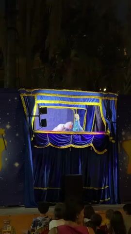 A puppet show is taking place at night. The stage is decorated with blue curtains and yellow trim, and has a backdrop of a starry sky with yellow stars. In the puppet stage, characters resembling Elsa and Olaf from Disney's Frozen are visible. In front of the stage, several children are seated, facing the performance. The lighting suggests an outdoor setting, possibly a park or public square, with palm trees visible in the background.