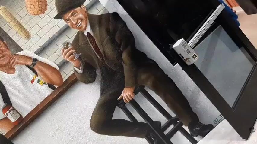 The video shows a mural outside of a restaurant. The mural depicts a scene of a deli counter.  There are portraits of a man in a suit holding a cat, another man in a white shirt, and Frank Sinatra.  Hanging from the awning are illustrations of meats.  The sign above reads "Mickey's Italian."  A man wearing a yellow head wrap is visible in the reflection of the glass in front of the mural.