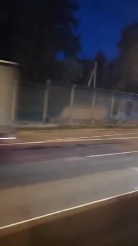 The video is a shaky, first-person perspective recording from a vehicle moving at night. The camera pans across a road with streetlights and trees visible. Several cars are seen on the road, some of which are blurred due to motion. The environment appears to be a suburban or rural area with a dark sky overhead. There is a bus stop with a shelter visible in the distance. The video focuses on the movement of vehicles and the ambient light of the streetlamps.