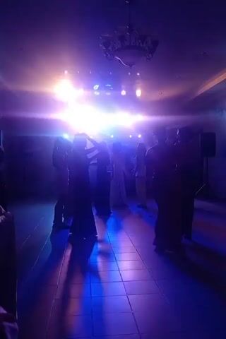 A group of women in formal attire are dancing in a dimly lit room illuminated by bright blue and white stage lights. The floor is tiled and reflective, showing colorful light patterns. The women are silhouetted against the lights, making their features indistinct. Some of them appear to be holding up their hands or adjusting their hair. A chandelier is visible hanging from the ceiling. The overall atmosphere suggests a celebratory event like a wedding reception or a formal party.