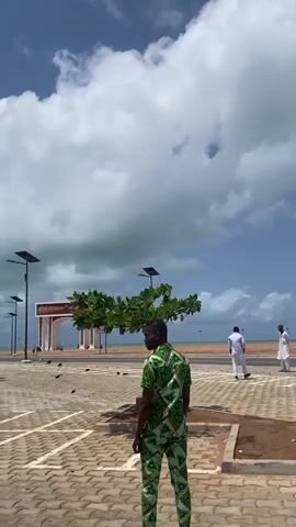 The video shows a man in a green, patterned suit standing in a brick-paved parking lot. In the distance, two men in white clothing walk away from the camera toward a light-colored beach and ocean under a partly cloudy sky. The parking lot has small trees, each equipped with a solar panel. Beyond the beach is a tan-colored wall with a decorative archway. Some people can be seen milling about in the distance.