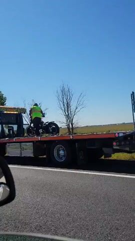 A tow truck is on the side of the road. A person in a neon vest is standing next to a motorcycle on the back of the tow truck. The tow truck has a logo that says "Logan" on it. There is a traffic cone on the road in front of the tow truck. The background is a field of grass and trees under a clear blue sky.