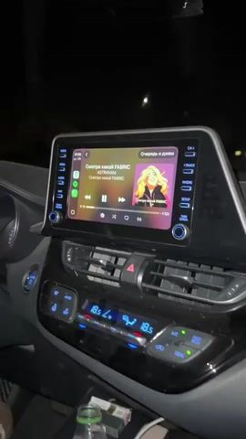 The video shows the interior of a car at night, focusing on the infotainment system playing music and then shifting to the driver, a young woman with long brown hair. The car’s dashboard is visible, with the infotainment screen displaying a song by ASTRXNXMX called "Смотка какой FABRIC" playing on Spotify. The car's temperature controls are set to 18.5 degrees Celsius. The camera moves to the driver, showing her profile as she drives. She smiles and appears to be enjoying the music, moving slightly with the beat. Raindrops are visible on the car window.