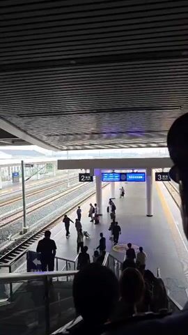 The video shows a busy train station platform with several people waiting and moving about. There are train tracks visible on the left, with overhead power lines. The platform itself is marked with yellow lines indicating the edge. A digital display board shows "2" and some Chinese characters, likely indicating a platform number. Several people are walking with luggage, and some are standing or sitting. An escalator is visible on the left side of the frame, with people descending. The overall environment appears to be an indoor or covered train station. The camera seems to be positioned on an upper level, looking down onto the platform.