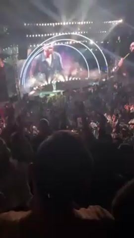 The video shows a concert with a large crowd and a performer on stage. The stage has large video screens displaying the performer. The crowd is dense and people are raising their hands and cheering. There are stage lights shining down on the crowd. The video is shot from within the crowd, looking towards the stage.