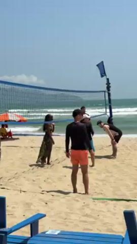 A group of six people is playing volleyball on a beach on a sunny day.  A woman in a floral dress stands to the left of a volleyball net, while several men wearing beach attire are positioned on the opposite side of the net.  The volleyball is in the air, having just been hit. Behind the players, the ocean stretches to the horizon with small waves visible. To the left of the scene, partially visible, are blue wooden beach chairs and an orange beach umbrella. A blue flag hangs from a pole near the volleyball net. The sky is light blue and mostly clear. The sand is light tan.