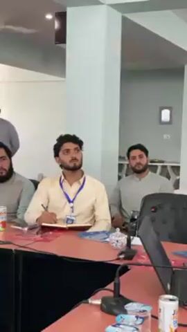 The video shows a group of approximately six to seven men attending what appears to be a meeting or presentation in an indoor conference room. The men are mostly young adults, seated around a long, reddish-brown rectangular table.

In the foreground, a man with a beard wearing a light yellow shirt and an ID badge around his neck is actively taking notes in a notebook with a pen. He is looking upwards, seemingly listening intently to a speaker who is not visible in the frame. To his left, another man with a beard is seated, also looking forward. Various drink cans and glasses are placed on the table in front of them, along with papers and microphones.

Further down the table to the left, two other men are seated, one wearing a dark blue shirt and glasses, and another in a blue and white patterned shirt, also with glasses. Both are looking towards the front, appearing engaged. Another man in a light gray shalwar kameez is visible seated to the right of the man in the yellow shirt. In the background, a man with a beard, dressed in a light gray traditional outfit, is standing.

The room has light-colored walls and pillars, with ceiling fans and windows fitted with blinds. A floral arrangement with white flowers is placed on the table. The lighting suggests it is daytime. The overall atmosphere appears focused and attentive, with participants listening and some taking notes. The scene is set in what looks like a professional or educational environment in Shurandam, Afghanistan.