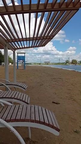 A sandy beach scene unfolds under a wooden pergola. In the foreground, three empty wooden lounge chairs face the water. Beyond the chairs, the sandy expanse of the beach stretches towards a calm body of water, possibly a lake or river. A blue lifeguard tower stands in the distance on the left. The sky is a clear blue with scattered white clouds. A few distant structures and trees line the far bank of the water. Towards the right of the frame, two figures, a man and a woman, walk near the water's edge. The overall atmosphere is peaceful and serene.