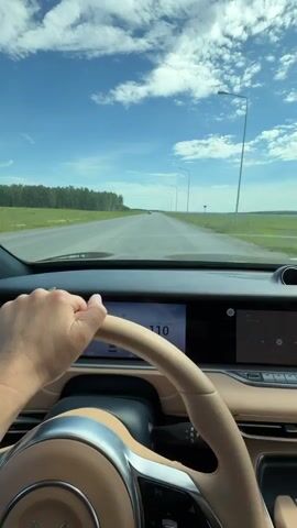 This is a first-person perspective video of someone driving a car on a sunny day. The driver's hands are visible on the steering wheel. The car's dashboard features a digital display with navigation information. Outside the car, the scene is a straight, paved road stretching into the distance, flanked by green fields and a line of trees on the left. The sky is blue with scattered white clouds. Several streetlights are visible along the side of the road. In the distance, other cars can be seen on the road. The overall mood is peaceful and serene, suggesting a leisurely drive. The time of day appears to be late morning or early afternoon, given the bright sunlight and clear skies. The location is identified as Severny, Russia. There is no visible text in the video itself.