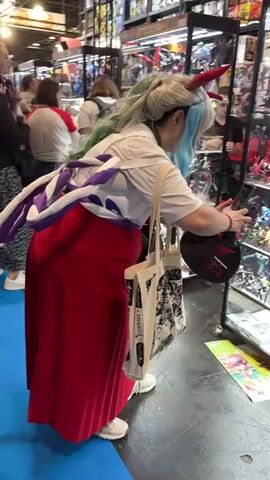 The video shows the interior of a store that appears to be a retail establishment selling anime and collectible merchandise. The store is well-stocked with shelves and display cases filled with various action figures, models, and other merchandise. The lighting is bright, illuminating the products.

In the foreground, a person is visible from behind, wearing a costume that includes a white shirt, a red skirt, and a long ponytail with light green and blue streaks. They are also wearing a white and purple sash or scarf draped over their shoulders and back. The person has a red horn on their forehead and is carrying a white tote bag with black illustrations. They are looking into a glass display case filled with merchandise.

To the left of this person, another individual wearing a black hooded garment and a patterned skirt is also looking into a display case. They are carrying a black backpack with a cartoon character attached.

In the background, other people can be seen browsing the store. The shelves are packed with boxed merchandise, mostly featuring anime characters and designs. The overall environment suggests a busy store, possibly during a convention or a weekend, with customers actively engaged in shopping. The floor is a dark color, possibly a dark blue or black. There are also some red boxes or storage containers visible on lower shelves to the right.