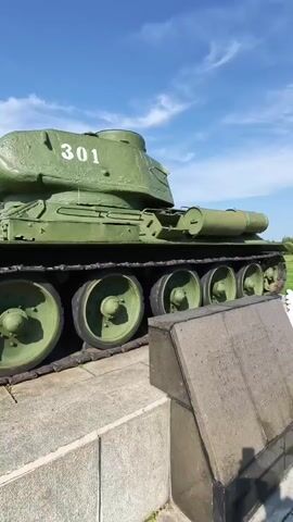 A static shot of a green Soviet T-34 tank on display. The tank is positioned on a concrete pedestal under a clear blue sky with a few white clouds. The side of the tank displays the number 301 in white. A memorial plaque is visible in the foreground to the right of the tank. The tank's tracks and road wheels are clearly visible. The image is captured from a low angle, looking slightly upwards.