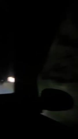 The video shows a night time view from inside a car. The car is driving on a road surrounded by trees. The headlights of the car illuminate the road ahead, and the surrounding darkness. There are other lights visible in the distance. A person's hands are visible on the steering wheel.