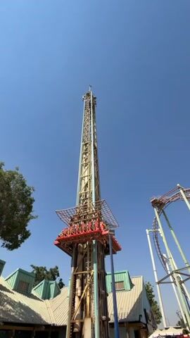 A drop tower amusement ride is shown against a clear blue sky.  The ride is tall and metallic, with a cage-like structure at the top that is carrying passengers. The passengers appear to be seated and secured. The ride mechanism is visible on the tower's central support. In the background, a roller coaster track curves upwards. The ride is situated within an amusement park, with low-rise buildings with green roofs visible at the base of the tower and fencing around the attraction.