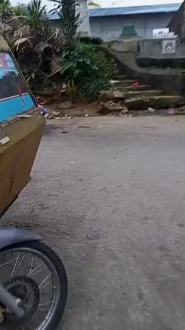 The video shows a street scene with several vehicles and piles of trash. In the foreground, a yellow and blue vehicle, possibly a tuk-tuk or a modified motorcycle, is partially visible on the left. The road surface is paved and appears somewhat worn. On the right side of the road, there is a steep embankment covered with dense vegetation and piles of debris, including what appears to be bamboo or dried plant material. Among the vegetation, there are several large piles of trash, consisting of plastic bags, fabric, and other refuse. Further up the embankment, there are steps leading to a building or structure with a blue roof and a white dome-like feature.

As the video progresses, several motorcycles pass by. One motorcycle, with a sidecar, is ridden by a person wearing a helmet and a dark jacket. Another motorcycle, with two people on it, approaches from the opposite direction. The person in front is wearing a blue shirt, and the person behind is wearing a dark shirt. Further down the road, a black pickup truck with the Mitsubishi logo is parked on the left. More trash is visible near the truck and the embankment. The overall environment appears to be a rural or semi-urban area with a clear indication of waste management issues. The sky is overcast, suggesting possibly cloudy weather.
